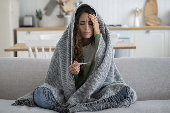 Mujer sentada y tapada con manta que sufre escalofríos sin fiebre mirando el termómetro.